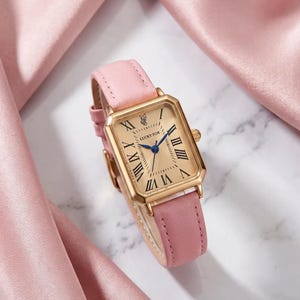 Puede incluir: Un reloj de pulsera rectangular con caja dorada y correa de cuero rosa claro. La esfera tiene fondo dorado, números romanos negros, manecillas azules y el texto LUCKY FOX con un pequeño icono de zorro en la parte superior central.