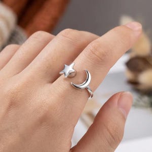 Puede incluir: Un anillo abierto de tono plateado que presenta un dije de estrella pulida en un extremo y un dije de media luna en el otro. La banda es lisa y metálica, diseñada con un espacio ajustable entre los dos motivos celestiales.