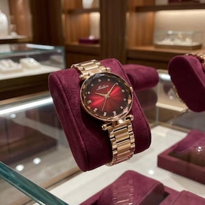 May include: A rose gold-tone wristwatch with a round, deep red sunray dial featuring the brand name Meibin in white script. The dial is accented with small, clear crystal hour markers and gold-tone hands, attached to a matching metal link band.