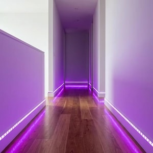 Può includere: Un corridoio con illuminazione a strisce LED viola brillante installata lungo i battiscopa. La luce proietta un vibrante bagliore violetto sul pavimento in legno lucido e sulle pareti bianche, creando un distinto effetto di illuminazione.