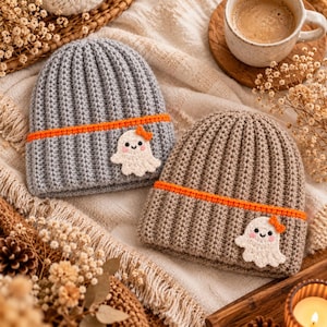 Könnte beinhalten: Zwei gerippte Häkelmützen, eine graue und eine beige, jeweils mit einem leuchtend orangefarbenen Querstreifen und einer kleinen weißen Geisterapplikation mit einer winzigen orangefarbenen Schleife und rosa Wangen aus strukturiertem Garn.