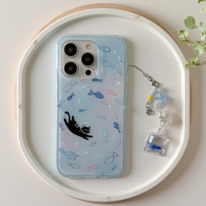 May include: A light blue smartphone case featuring a black cat illustration and various small fish outlines. The case includes a dangling charm attachment with a miniature clear plastic bag containing a tiny blue fish and a blue star bead.