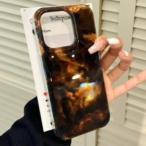 May include: A rectangular smartphone case featuring a glossy, dark brown and amber tortoiseshell pattern with swirling highlights. The case has a large cutout for a camera module and is displayed against a white card with Instagram text.
