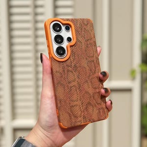 May include: A rectangular smartphone case featuring a brown snakeskin-patterned back panel and a bright orange frame. The camera cutout area is also outlined in orange, surrounding the multiple circular lenses of the mobile device.