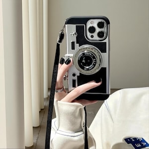 May include: A black and silver phone case designed to resemble a vintage camera, featuring a prominent circular lens element in the center, a rectangular body shape, and a black fabric lanyard strap attached to the side for carrying convenience.