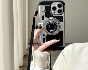 May include: A black and silver phone case designed to resemble a vintage camera, featuring a prominent circular lens element in the center, a rectangular body shape, and a black fabric lanyard strap attached to the side for carrying convenience.