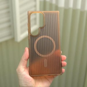 May include: A translucent brown smartphone case featuring a vertical ribbed texture across the back surface. The case includes a large cutout for a camera array at the top left and a circular magnetic charging ring centered on the back panel.