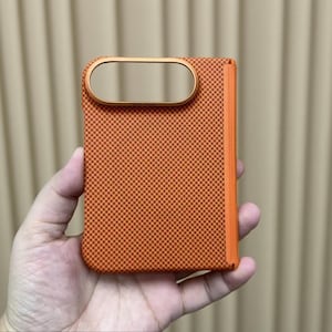 Peut inclure: Une coque de téléphone rectangulaire à motif damier orange et marron foncé avec une tranche orange lisse. La coque présente une découpe horizontale ovale pour l'appareil photo, encadrée par un bord orange métallique avec la marque Sulada.
