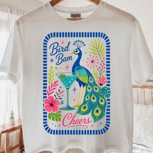 May include: A white t-shirt featuring a graphic print of a peacock holding a martini glass. The design includes the text Bird Bam and Cheers, surrounded by a blue and white striped rectangular border with colorful tropical floral accents.