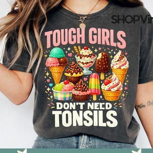 May include: A charcoal grey t-shirt featuring the text TOUGH GIRLS DON'T NEED TONSILS in pink and white lettering. The graphic includes colourful illustrations of various ice cream cones, sundaes, cupcakes, and a popsicle surrounded by small hearts.