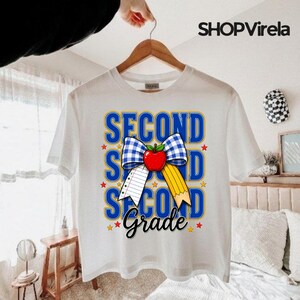 Può includere: Una t-shirt bianca a maniche corte con una grafica che riporta il testo "SECOND GRADE" in lettere blu. Il design include un fiocco a quadretti blu e bianco, una mela rossa, un foglio di carta a righe, una matita gialla e piccole stelle rosse.