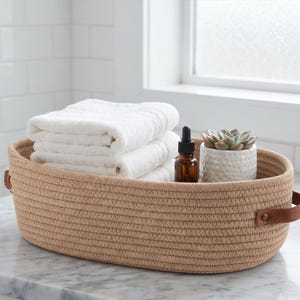 May include: An oval-shaped storage basket made of tan coiled rope with brown leather handles on each side. Inside the basket are two folded white towels, a small amber glass dropper bottle, and a white ceramic pot containing a green succulent.