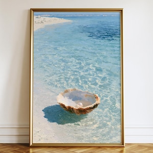 May include: A framed vertical wall art print depicting a single white pearl resting inside an open textured seashell. The shell sits in shallow, clear turquoise water near a sandy white beach under a bright blue sky, captured in a realistic style.