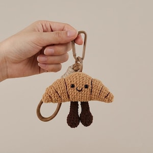 May include: A tan crocheted croissant keychain featuring a smiling face with black bead eyes and a stitched mouth. The croissant has two dark brown crocheted legs dangling from the bottom and is attached to a tan cord with a brown plastic clip.