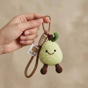Può includere: Un portachiavi a forma di pera lavorato all'uncinetto in verde chiaro con un viso sorridente, piedini marroni e una piccola foglia verde sulla parte superiore. Presenta un cordino marrone, un moschettone in metallo e un'etichetta in tessuto.