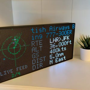 May include: A rectangular black LED pixel display board showing flight data including British Airways B, 777-300ER, LHR to JFK route, 36,000ft altitude, 480kts ground speed, 5.0nm distance, and North East direction with a green radar graphic.