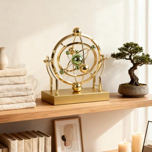 May include: A kinetic sculpture featuring a gold-toned metal frame with rotating rings and spheres. The central structure includes a large green sphere surrounded by smaller gold and green spheres, all mounted on a rectangular gold base.