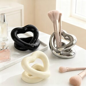 May include: Three heart-shaped holders in black, silver, and cream finishes. The silver holder contains two makeup brushes with light pink bristles. A perfume bottle is visible nearby, and two additional makeup brushes lie on the surface.