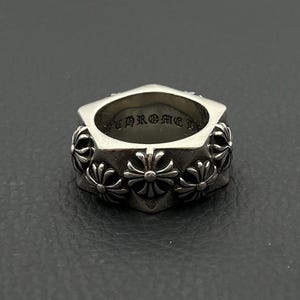 Pode incluir: Um anel de metal prateado com uma banda exterior hexagonal decorada com múltiplos motivos de cruz em relevo. O interior da banda está gravado com o texto CHROME HEARTS numa fonte distinta de estilo gótico, visível ao longo da borda.