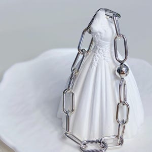 May include: A silver chain bracelet featuring a mix of smooth, polished rectangular links and textured, snake-chain style links. The bracelet includes a single spherical bead with visible lettering, arranged in a continuous loop design.