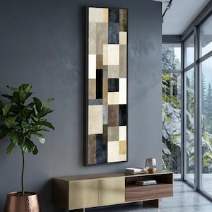 May include: A tall, vertical rectangular wall art piece featuring a geometric composition of stacked, multi-sized blocks in shades of cream, beige, tan, dark brown, charcoal gray, and black, all enclosed within a thin, dark-colored frame.