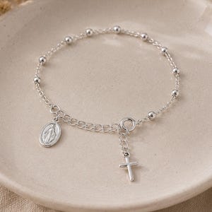 May include: A silver chain bracelet featuring small round beads, a circular Miraculous Medal charm, and a small dangling cross pendant. The bracelet is constructed with a delicate link chain and a circular clasp, showcasing a polished metallic finish.
