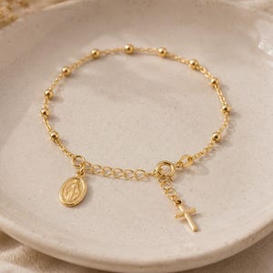 Puede incluir: Una pulsera de cadena en tono dorado con pequeñas cuentas redondas espaciadas a lo largo. La pulsera incluye un cierre de resorte circular, un pequeño dije de cruz colgante y un colgante ovalado que representa una figura en relieve.