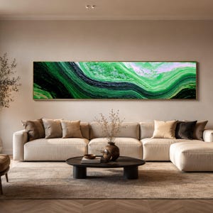 Puede incluir: Una pieza de arte mural rectangular horizontal y alargada que presenta una pintura abstracta fluida con capas arremolinadas de tonos verde bosque profundo, esmeralda, negro y blanco, en un marco dorado fino sobre un sofá beige.