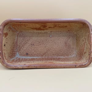 May include: A rectangular ceramic loaf pan with a speckled tan and brown glaze. The vessel features two integrated handles on the short ends, each decorated with a raised, curved, and ribbed pattern along the outer edges of the rim.