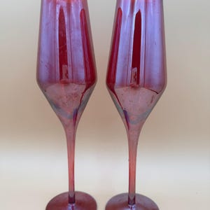 May include: A pair of tall, slender champagne flutes crafted from translucent red glass. Each glass features a smooth, elongated bowl that tapers into a thin, delicate stem, resting on a circular base, all finished with a consistent deep red hue.