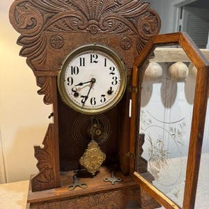 May include: A wooden mantel clock with an ornate carved top and a white circular face featuring black Arabic numerals. The clock has a hinged glass door with etched floral designs, a brass pendulum, and two metal winding keys on the base.