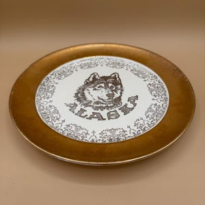 May include: A round decorative plate with a metallic gold rim and a white center. The center features a brown illustration of a wolf head above the text ALASKA, surrounded by a detailed brown floral pattern that encircles the central design.