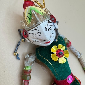 Könnte beinhalten: Eine handgefertigte Holzpuppe mit weiß bemaltem Gesicht, schwarzen Liniendetails, roten Lippen und einem strukturierten Kopfschmuck. Die Figur trägt ein dunkelgrünes Stoffoberteil, das mit einer gelben Metallblume und einer roten Paillette verziert ist, aufgehängt an einer goldenen Kordel.