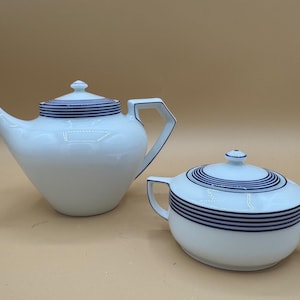 May include: A white porcelain teapot and a matching lidded sugar bowl, both featuring a series of thin, dark blue horizontal stripes around the lids and the upper bodies. The teapot has a curved spout and an angular handle, while the sugar bowl has two side handles.