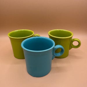 Könnte beinhalten: Drei Keramikbecher in einer Gruppe angeordnet. Ein Becher ist in leuchtendem Türkisblau gehalten, während die anderen beiden in hellem Limettengrün sind. Jeder Becher hat eine glatte, glänzende Oberfläche, einen zylindrischen Körper und einen kleinen Henkel.