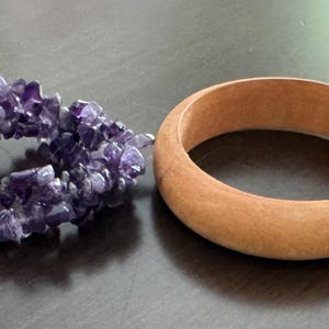 May include: Four bracelets arranged in a row: a wide, smooth, light brown resin bangle; a cluster of raw, deep purple amethyst chips on an elastic cord with a small white circular tag; a smooth, medium-toned wooden bangle; and a textured wooden slat bracelet.