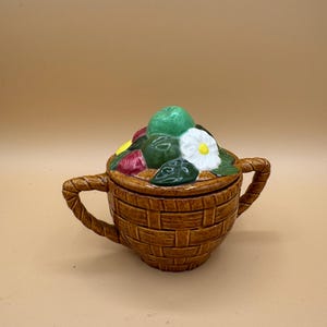May include: A brown ceramic sugar bowl with a woven basket texture and two rope-style handles. The lid features a sculpted arrangement of a green apple, a red fruit, a yellow lemon, a white flower with a yellow centre, and green leaves.