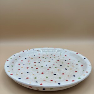 Könnte beinhalten: Ein runder Keramikt runder Keramikteller mit weiß glasierter Oberfläche, der mit einem verstreuten Muster aus kleinen, bunten Punkten in Rot, Blau und Grün verziert ist. Der Teller hat einen leicht erhöhten Rand und eine glatte, glänzende Oberfläche.