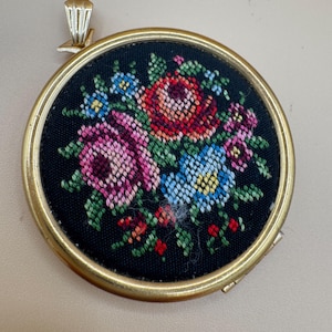 May include: A round pendant featuring a black fabric insert with a cross-stitch floral pattern in shades of red, pink, and blue. The embroidery is encased in a gold-toned metal frame with a matching bail at the top for attachment to a chain.
