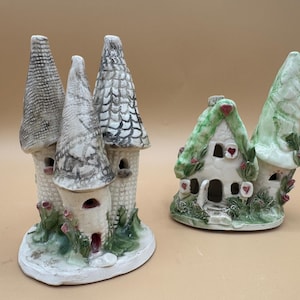 Puede incluir: Dos estructuras en miniatura de cerámica. La pieza de la izquierda presenta tres torres altas con techos grises texturizados, pequeñas aberturas arqueadas y follaje verde en la base. La pieza de la derecha es una casita blanca con techo verde y corazones rojos.