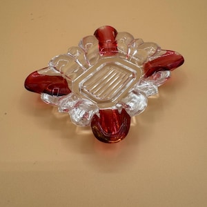 May include: A square glass ashtray featuring a clear center with a ribbed base and four rounded corners accented with deep red glass. The design includes four distinct indentations along the edges, creating a symmetrical and functional glass piece.