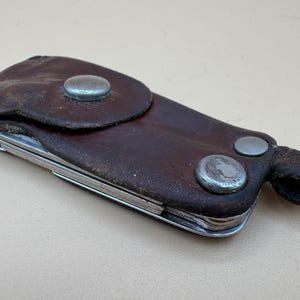 Op de afbeelding: Een donkerbruine lederen sleuteletui met een overslagflap die wordt vastgezet met een zilverkleurige metalen drukknoop. De etui bevat twee extra zilverkleurige metalen klinknagels en een kleine lederen lus aan de zijkant voor bevestiging.