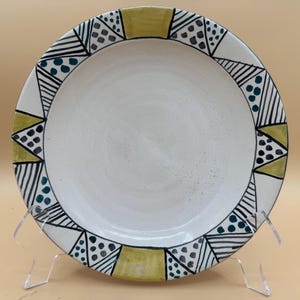 Puede incluir: Un plato redondo de cerámica con un centro blanco liso y un borde decorativo que presenta patrones geométricos alternos en tinta negra, puntos verde azulado y bloques de color verde oliva sólido. El plato está sobre un soporte de plástico.