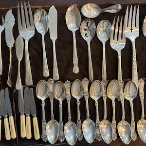 May include: A collection of silver-toned flatware including various forks, spoons, and knives. Several knives feature cream-colored handles with the word LONDON printed on them. The set is arranged on a dark brown fabric surface with individual slots.