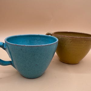 May include: Two ceramic mugs with a rounded shape and a smooth, glazed finish. One mug is a vibrant turquoise blue with a curved handle, while the second mug is a muted mustard yellow. Both items feature a simple, wide-rimmed design.