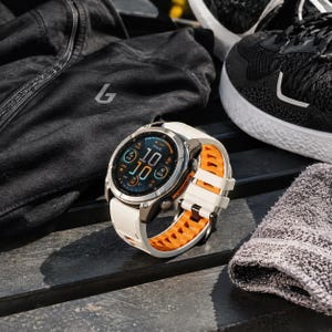 May include: A Garmin Fenix smartwatch featuring a round digital display showing the time 10:10, a metallic bezel, and a two-tone silicone watch band with a white exterior and a bright orange interior with a textured, ventilated design.