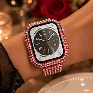 Könnte beinhalten: Eine Smartwatch mit rechteckigem Gehäuse, schwarzem Analog-Zifferblatt und einem metallisch roten Schutzrahmen, der mit zahlreichen klaren, funkelnden Strasssteinen besetzt ist. Das passende rote Armband ist ebenfalls mit kleinen Kristallen bedeckt.