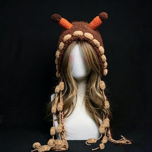 May include: A brown fuzzy crochet hat featuring two orange and brown antennae on top, a border of small beige pom-poms around the face opening, and long braided side tassels adorned with additional beige pom-poms hanging down the sides.
