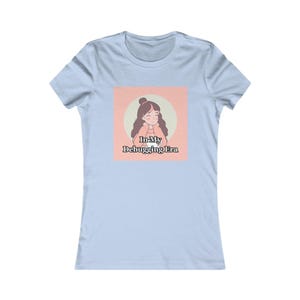 May include: A light blue short-sleeve t-shirt featuring a square graphic on the chest. The graphic shows an illustration of a person with brown hair in a bun, wearing a peach sweater, holding a white mug, with the text In My Debugging Era.