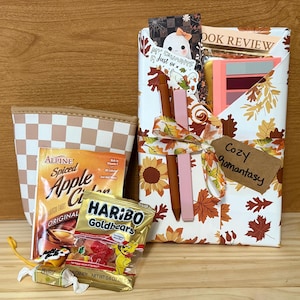 Puede incluir: Un set de regalo que incluye un estuche a cuadros, un paquete de sidra de manzana especiada Alpine, una bolsa de Haribo Goldbears, un caramelo y un libro envuelto en papel blanco con hojas de otoño y girasoles, atado con una cinta y una etiqueta Cozy Romantasy.